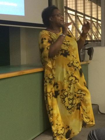 Na foto, a palestrante Esmeralda Ribeiro, durante a palestra, gesticulando. Ela é uma mulher negra de pele escura, cabelos curtos e grisalhos, usa óculos e veste um vestido longo amarelo, relógio e sapatilhas brancas.