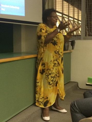 Na foto, a palestrante Esmeralda Ribeiro, durante a palestra, gesticulando. Ela é uma mulher negra de pele escura, cabelos curtos e grisalhos, usa óculos e veste um vestido longo amarelo, relógio e sapatilhas brancas.